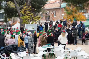 Un grupo de personas, algunas con gorros de duende rojos y verdes, se reúnen al aire libre en una plaza con sillas blancas y árboles. La escena, como un animado "borrador automático", muestra a personas de pie o sentadas entre edificios y follaje otoñal.