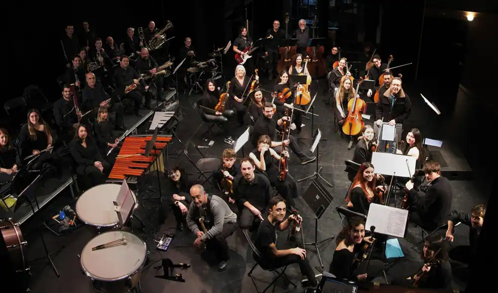 La Orquesta Athanor está sentada y de pie en un escenario oscuro de Rivas, con cuerdas, metales y percusión. Los músicos están colocados por secciones, de cara a la cámara y mirando hacia arriba, listos para interpretar el Carnaval de los animales.