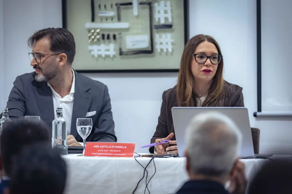 En el Rivas Business Meet Up, dos personas se sientan a una mesa durante una reunión formal en la ciudad. La mujer de la derecha habla con un ordenador portátil y una placa de identificación delante, mientras los miembros del público observan este notable evento de polo de inversión.