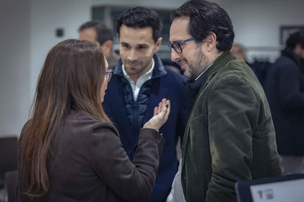 Una mujer gesticula mientras habla con dos hombres en el Rivas Business Meet Up. Los tres, vestidos con chaqueta, parecen enzarzados en una conversación sobre la ciudad y su papel como polo de inversión, con otras personas difuminadas al fondo.