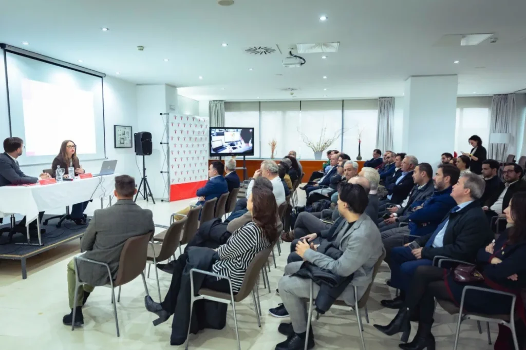 En el Rivas Business Meet Up, un grupo vestido con traje de negocios se sienta en filas frente a una pantalla de presentación. Dos oradores con micrófonos dirigen el debate, mientras un asistente levanta la mano. Un monitor de TV muestra más información sobre la ciudad como polo de inversión.