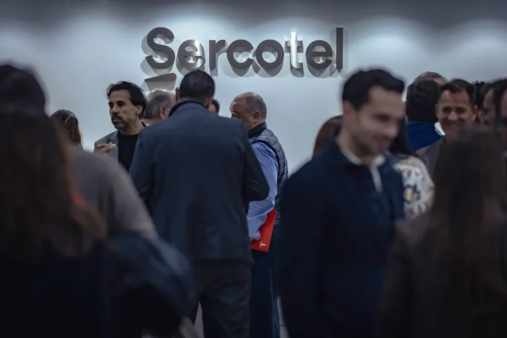 Un grupo de personas conversa en un abarrotado espacio interior bajo un gran cartel de Sercotel en el Rivas Business Meet Up, un dinámico evento de networking en la ciudad conocido como un emergente polo de inversión.