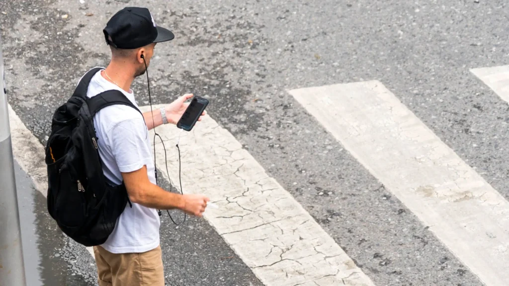 Una persona con gorra negra, camiseta blanca, pantalones cortos de color canela y mochila se para en un paso de peatones con un teléfono en la mano y auriculares en los oídos, mirando hacia la carretera, dispuesta a cruzar calles donde el pavimento desgastado marca la guía de los peatones.