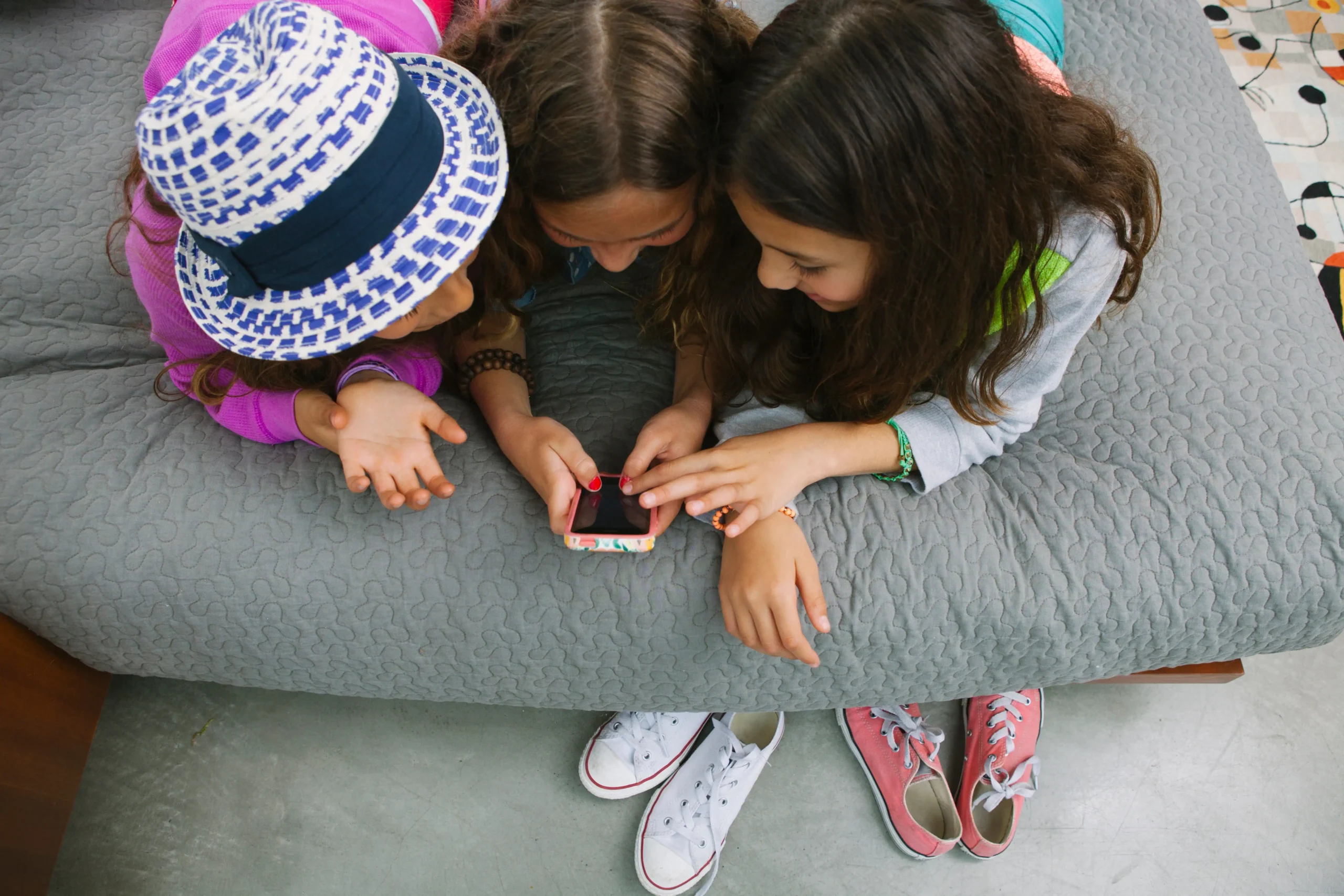 Tres niños de pelo largo están tumbados en una colcha acolchada gris, utilizando juntos un smartphone, lo que refleja el papel de la tecnología para menores. Dos pares de zapatillas descansan junto a la cama y un niño lleva un sombrero estampado.