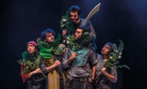 Durante el estreno en Rivas, cinco actores disfrazados se sitúan en el escenario sosteniendo grandes hojas verdes y plantas. Un actor, elevado sobre los demás, empuña una gran espada mientras todos adoptan poses expresivas y miran en distintas direcciones.