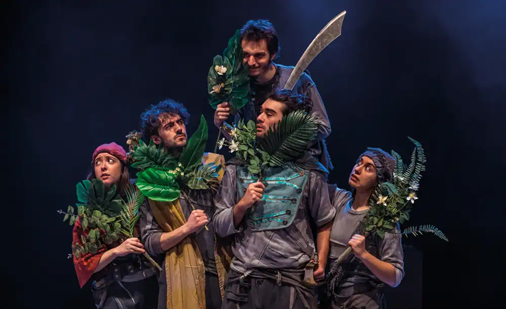 Durante el estreno en Rivas, cinco actores disfrazados se sitúan en el escenario sosteniendo grandes hojas verdes y plantas. Un actor, elevado sobre los demás, empuña una gran espada mientras todos adoptan poses expresivas y miran en distintas direcciones.