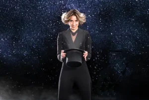 En el Festival de Magia de Rivas, una persona vestida de negro, sobre un fondo oscuro y estrellado, sostiene un sombrero de copa negro mientras partículas de nieve o confeti flotan en el aire, evocando la magia de la Navidad.