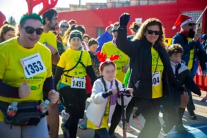 Un grupo de personas con camisetas amarillas y dorsales numerados participan en la carrera popular San Ripense en Rivas. Una mujer levanta el puño, mientras un niño con cuernos rojos de reno monta en patinete. Otros sonríen y caminan juntos.