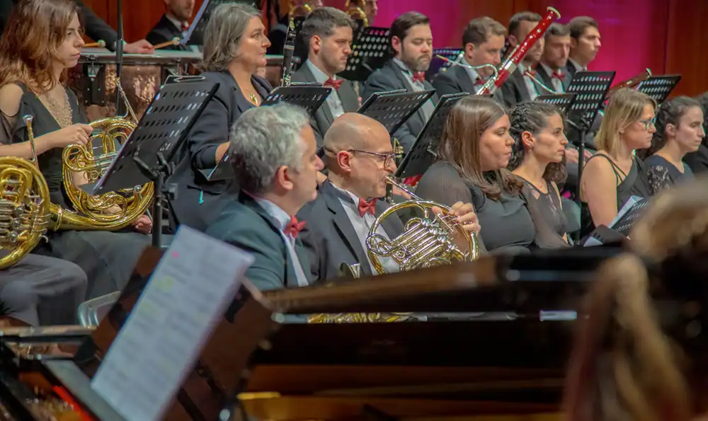 Músicos en traje de etiqueta actúan en la orquesta La Sinfónica de Rivas, tocando instrumentos de viento-metal y madera. Se ven atriles con partituras y, al fondo, más músicos e instrumentos en el Abono Sinfónico 2026.