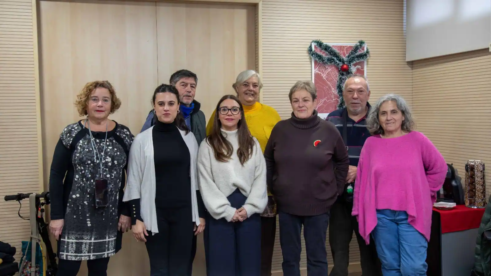 Ocho adultos están de pie en el interior, en fila india, mirando a la cámara. El ambiente parece informal, con una corona navideña en la pared detrás de ellos. Todos están vestidos con ropa de diario, lo que sugiere una reunión amistosa de Cohabita Rivas.