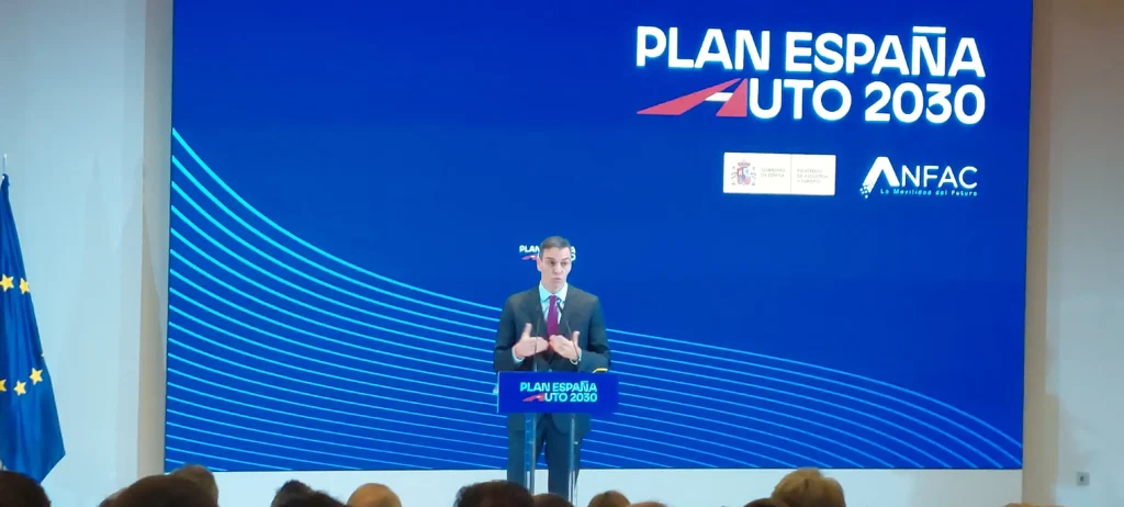 Una persona, de pie en un podio con el rótulo Plan España Auto 2030, se dirige al público para hablar de las ayudas directas y el despliegue de recargas. Detrás, una gran pantalla azul muestra el Plan España Auto 2030, logotipos de ANFAC y referencias a iniciativas relacionadas con el coche eléctrico.