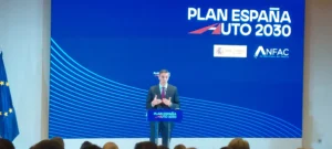 Una persona, de pie en un podio con el rótulo Plan España Auto 2030, se dirige al público para hablar de las ayudas directas y el despliegue de recargas. Detrás, una gran pantalla azul muestra el Plan España Auto 2030, logotipos de ANFAC y referencias a iniciativas relacionadas con el coche eléctrico.