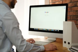 Una persona con camisa azul claro se sienta en un escritorio de madera con un iMac en el que se ve la página de inicio de Google y la función "borrador automático". El escritorio sostiene un teclado blanco, un ratón y dos organizadores de archivos blancos.
