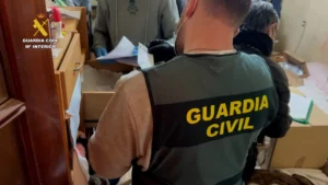 Un agente de la Guardia Civil ataviado con un chaleco en el que se lee Guardia Civil permanece de pie frente a un cajón abierto mientras otra persona manipula documentos en una sala durante una inspección relacionada con una red criminal de estafas online.