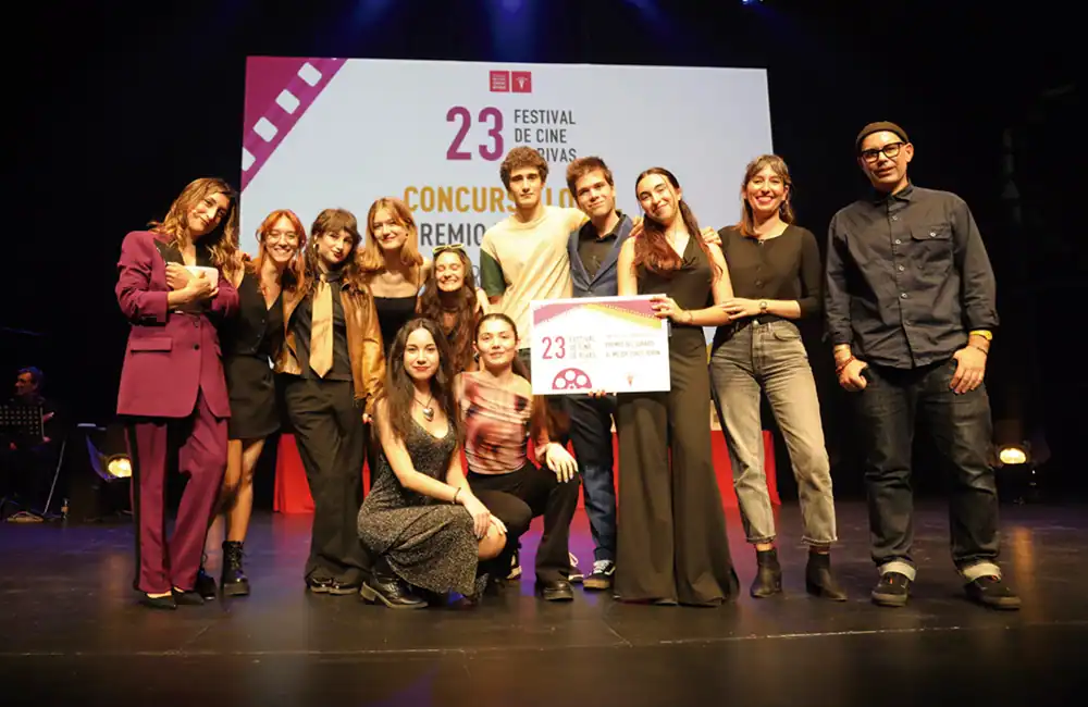 Un grupo de doce personas posa en el escenario, algunas de pie y otras arrodilladas, sosteniendo un gran certificado. Detrás de ellos, una pantalla muestra 23 Festival de Cine Rivas con texto adicional. El escenario parece ser un acto de entrega de premios de edición de vídeo o montaje. Un grupo de doce personas posa en el escenario, algunas de pie y otras arrodilladas, sosteniendo un gran certificado. Detrás de ellos, una pantalla muestra 23 Festival de Cine Rivas con texto adicional. El escenario parece ser un acto de entrega de premios de edición de vídeo o montaje.