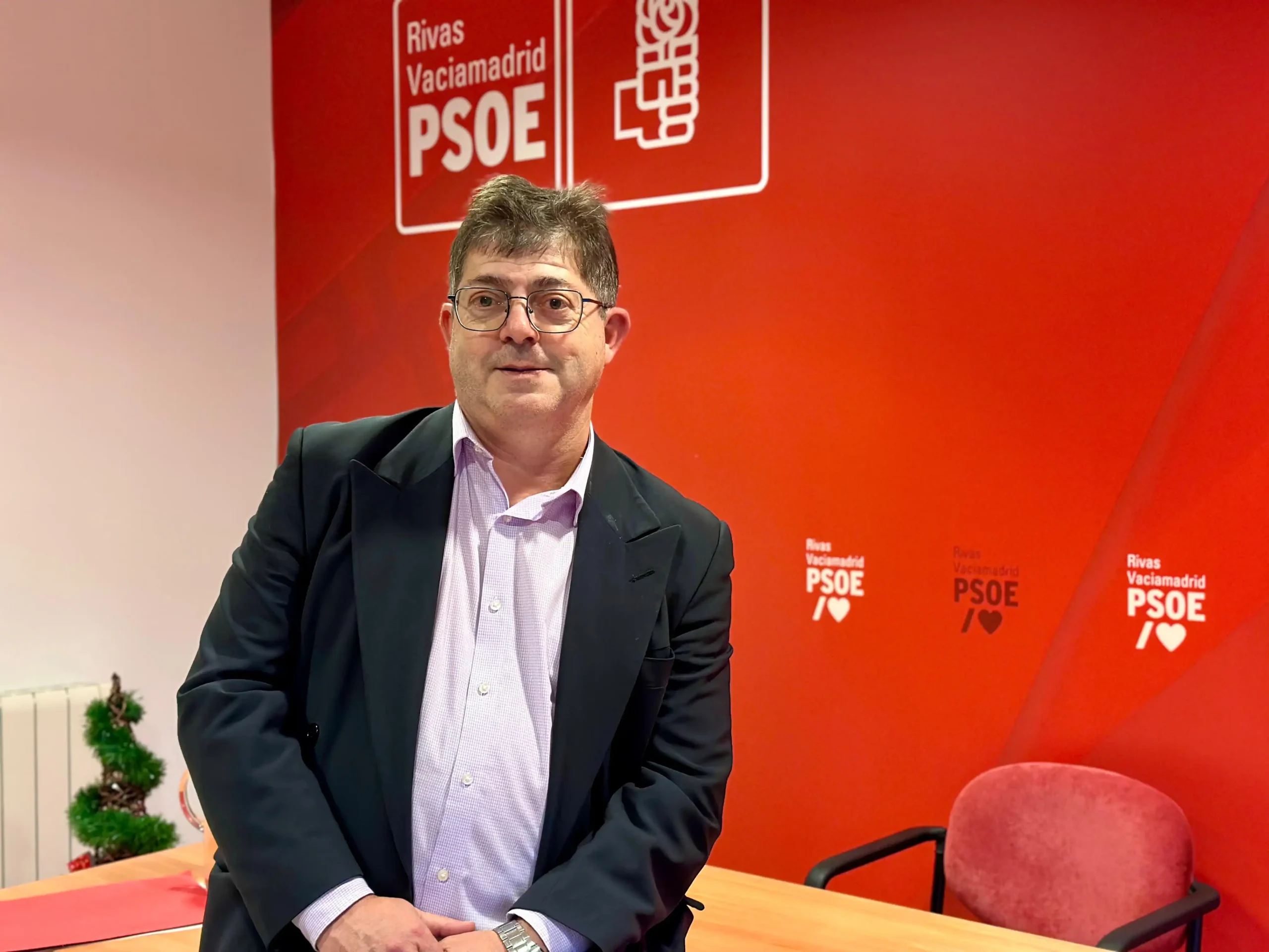 Alberto Cabeza, secretario general, con traje y gafas está de pie junto a una mesa en un despacho con paredes rojas que muestran los logotipos del PSOE de Rivas Vaciamadrid. Cerca, una silla roja y una pequeña planta completan la escena, en la que se pueden revisar documentos como un borrador automático.