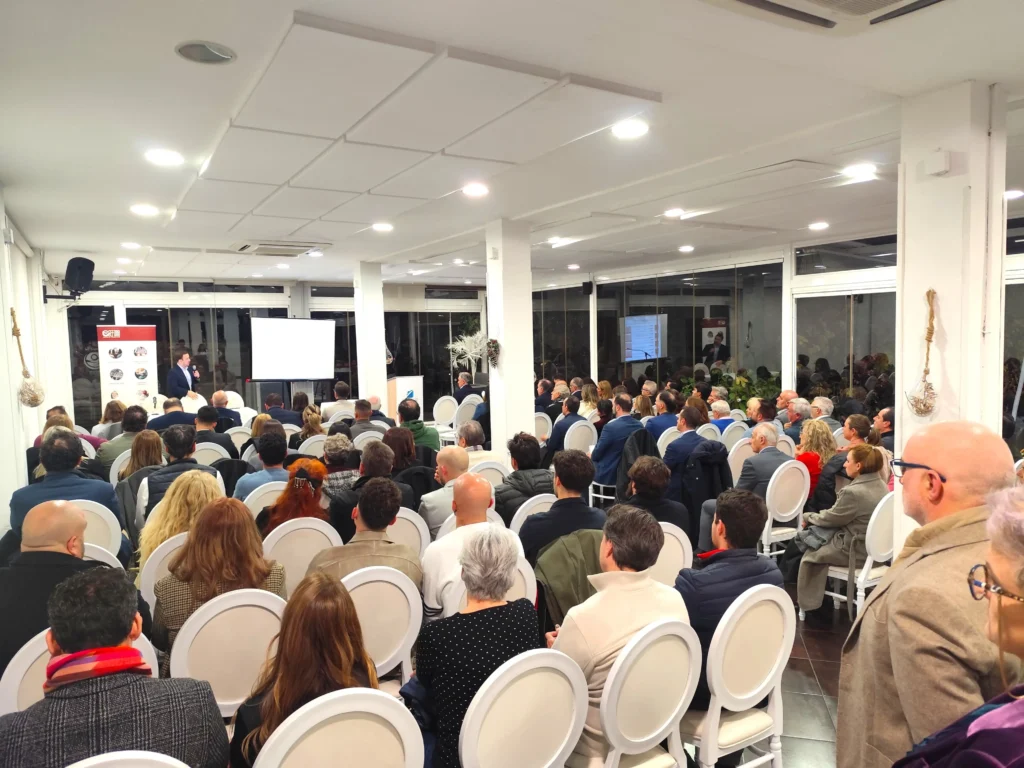 Un nutrido grupo de empresarios se sienta en sillas blancas en una sala de conferencias muy iluminada, frente a un escenario donde habla un presentador de ASEARCO cerca de una pantalla de proyección. La sala tiene paredes blancas, pilares y grandes ventanales durante el encuentro navideño.
