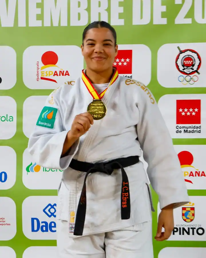 Nisrin Bousbaa, atleta de judo con gi blanco y cinturón negro, sostiene su medalla de oro y sonríe ante un telón de fondo con logotipos y texto en español tras ganar su tercer oro nacional.