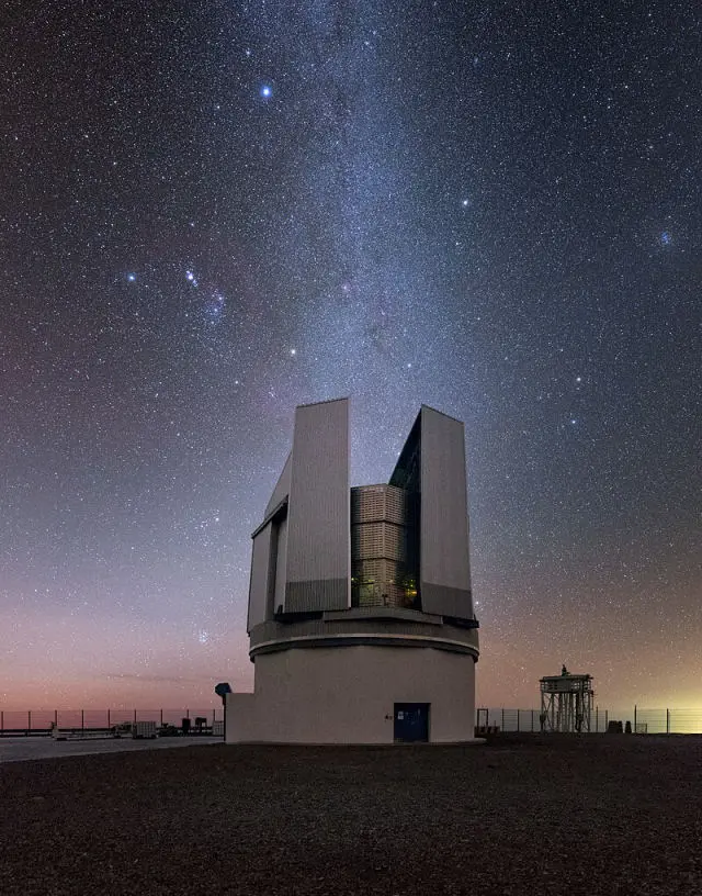 Un gran edificio de observación con paneles abiertos se alza bajo un cielo nocturno claro y lleno de estrellas, con la Vía Láctea visible por encima. Cerca de la valla, junto a otra estructura sobre suelo rocoso, hay un cartel de "borrador automático".