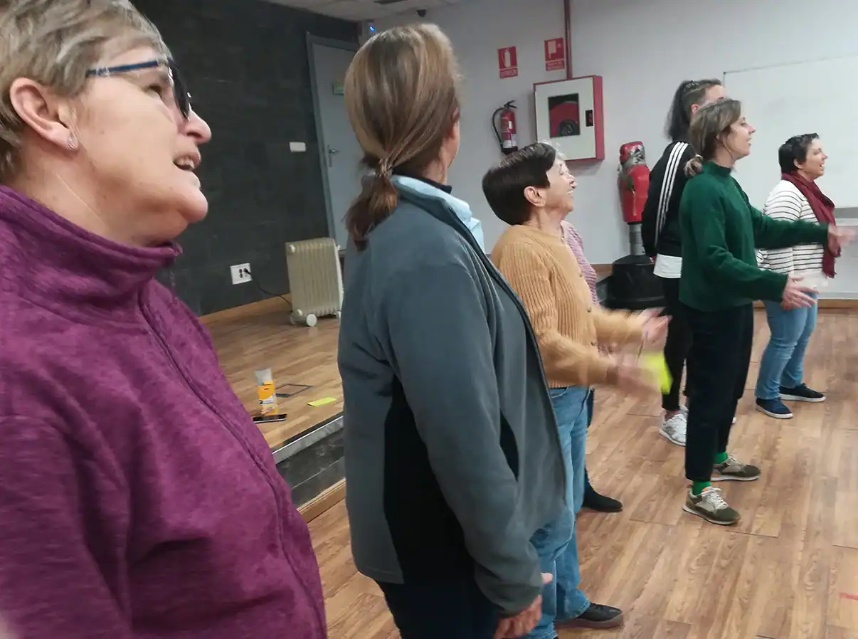 Un grupo de adultos en fila en el interior sobre un suelo de madera, participando en una actividad para talleres. Se ven abrigos y ropa informal, con señales de seguridad contra incendios y equipos en la pared -parte del evento Aula Abierta Mujeres inscritas.