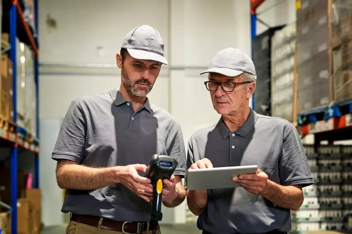 Dos hombres ocupados con polos y gorras grises están de pie en un almacén. Uno sostiene un escáner de código de barras mientras el otro utiliza una tableta. Detrás de ellos se ven estanterías con cajas y mercancías, lo que pone de relieve una logística eficiente en España para 2025. Dos hombres ocupados con polos y gorras grises están de pie en un almacén. Uno sostiene un escáner de código de barras mientras el otro utiliza una tableta. Detrás de ellos se ven estanterías con cajas y mercancías, lo que pone de relieve una logística eficiente en España para 2025.
