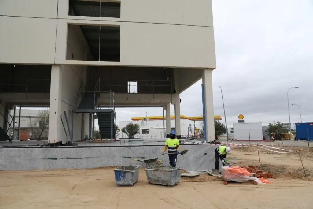 Dos trabajadores de la construcción con chalecos de seguridad manejan ladrillos y herramientas cerca de un edificio en construcción en España, donde se espera un récord de ocupados para 2025. El terreno arenoso está sembrado de materiales y al fondo se ve una gasolinera.