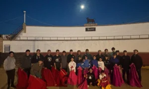 Un grupo de unas 25 personas, en su mayoría niños y adolescentes, posan juntos en una plaza de toros al aire libre al anochecer, cada uno con un capote rojo o rosa. Una luna y una estatua de un toro se ven en el borde superior del muro de la plaza.