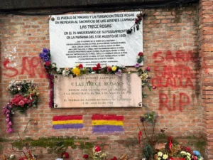 Un muro de ladrillo muestra dos placas y flores conmemorativas de Las Trece Rosas. Se ven pancartas, pintadas y amenazas rojas. Ambas placas -escritas en español- honran a las jóvenes ejecutadas en 1939 en la tumba de las 13 Rosas.