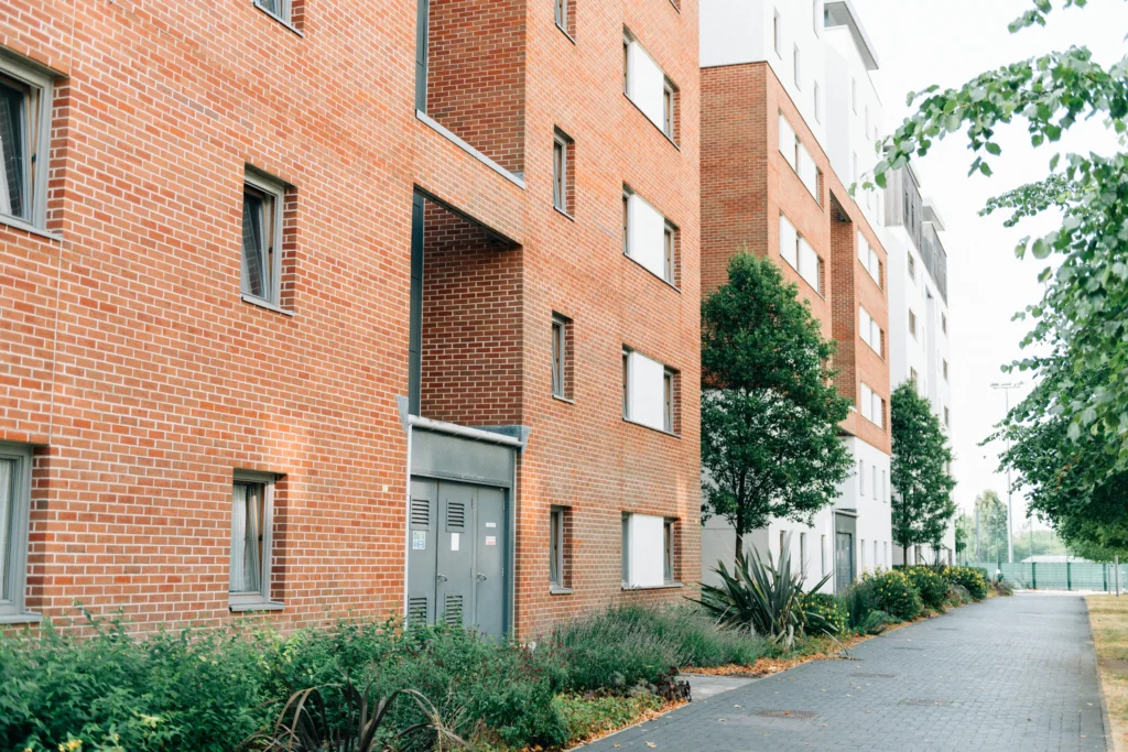 Se muestra un moderno edificio de la comunidad de vecinos con paredes de ladrillo rojo y ventanas rectangulares. Hay plantas y pequeños árboles a lo largo de una acera pavimentada junto al edificio en un día despejado.