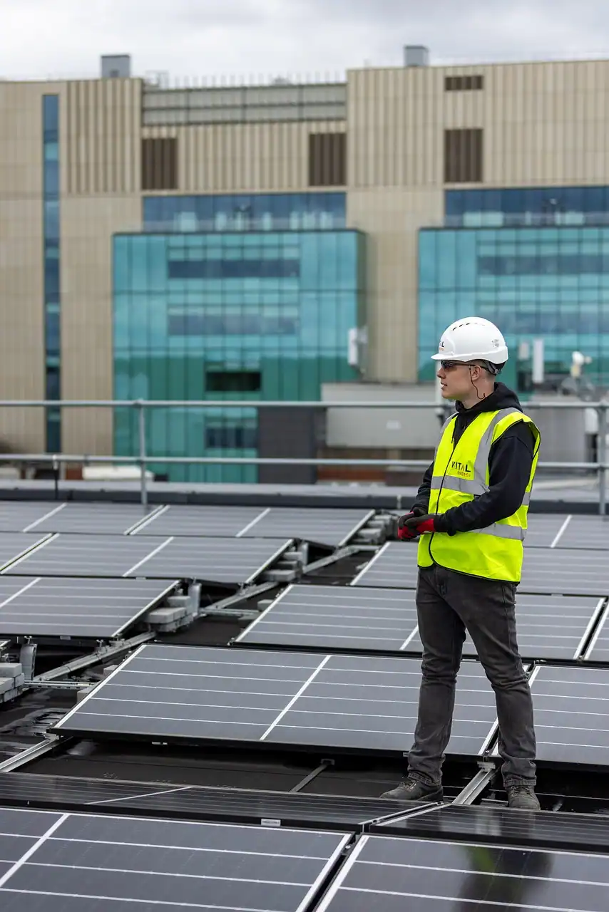 Una persona con casco blanco, gafas de seguridad y chaleco amarillo de alta visibilidad se encuentra entre paneles solares en un tejado, comprobando un borrador automático, con un moderno edificio al fondo.