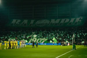 Los jugadores se alinean en un campo de fútbol antes de un partido de LA LIGA. En las gradas, los aficionados ondean banderas verdes y blancas y despliegan una gran pancarta en la que se lee BALOMPIÉ, creando un animado ambiente de estadio.