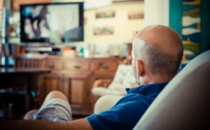 Un hombre calvo con camisa azul está sentado en el sofá de su salón, viendo la televisión, tal vez perdido en atracones de serie. El televisor aparece difuminado en el fondo, junto con muebles de madera y objetos decorativos.