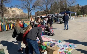 La gente se reúne al aire libre en la soleada Rivas, mirando los artículos expuestos para la venta o el intercambio. Algunos hacen cola, mientras otros interactúan con los productos. Al fondo se ven una valla de colores y varios edificios que contribuyen a crear un ambiente animado.