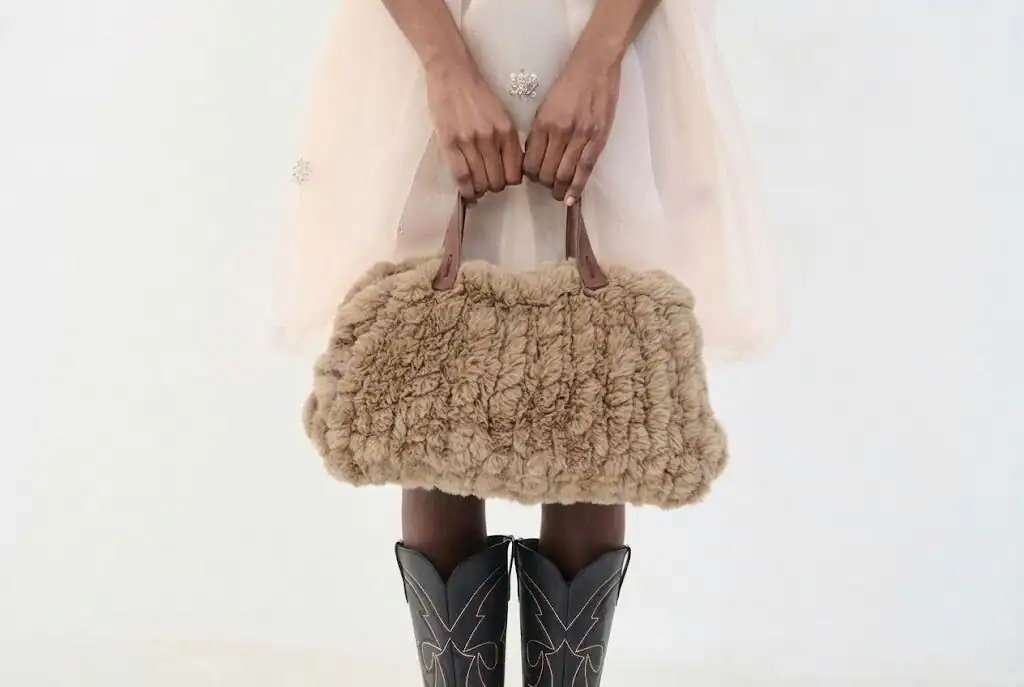 Una persona con una falda pálida y transparente sostiene un bolso de piel sintética marrón con asas marrones. Con botas vaqueras negras con pespuntes blancos, posan sobre un fondo blanco liso, imitando el estilo desenfadado de un borrador automático.