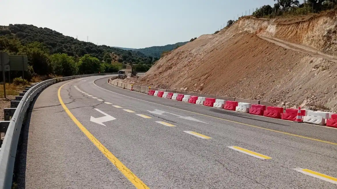 Una carretera de dos carriles traza una curva a la izquierda con claras marcas de carril amarillas y blancas, que recuerdan a un borrador automático. Barreras rojas y blancas bordean el lado derecho cerca de una ladera con suelo expuesto, mientras que al fondo se ven árboles y colinas.