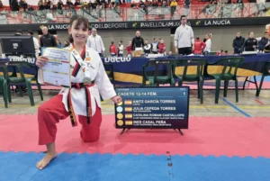 Un joven artista marcial de taekwondo de Rivas se arrodilla sobre una colchoneta roja y azul, sosteniendo con orgullo un certificado y mostrando medallas de oro Nacional y bronces Nacional. Un marcador muestra los nombres de los competidores mientras la gente se reúne en el fondo del recinto deportivo.