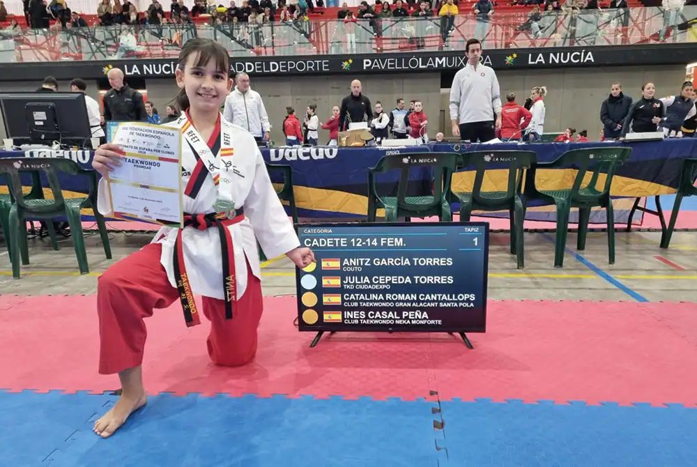 Un joven artista marcial de taekwondo de Rivas se arrodilla sobre una colchoneta roja y azul, sosteniendo con orgullo un certificado y mostrando medallas de oro Nacional y bronces Nacional. Un marcador muestra los nombres de los competidores mientras la gente se reúne en el fondo del recinto deportivo.