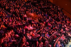 Un numeroso público sentado en filas de butacas en el interior de un teatro de Rivas asiste a un acto de la temporada cultural 2026. La iluminación tiñe de rojo a la multitud, la mayoría atenta, algunos mirando hacia el escenario y la dirección de Miguel del Arco.