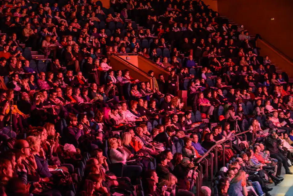 Un numeroso público sentado en filas de butacas en el interior de un teatro de Rivas asiste a un acto de la temporada cultural 2026. La iluminación tiñe de rojo a la multitud, la mayoría atenta, algunos mirando hacia el escenario y la dirección de Miguel del Arco.