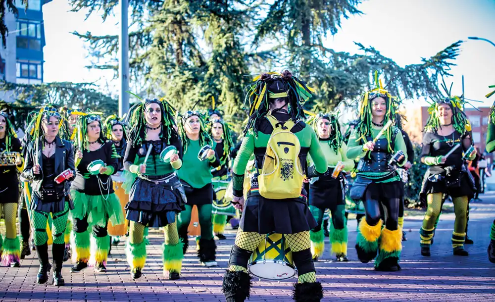 Un grupo de personas vestidas con trajes verdes, negros y amarillos con accesorios a juego tocan tambores e instrumentos de percusión al aire libre en el Carnaval 2026 Rivas, de pie sobre una superficie de ladrillo con árboles al fondo.