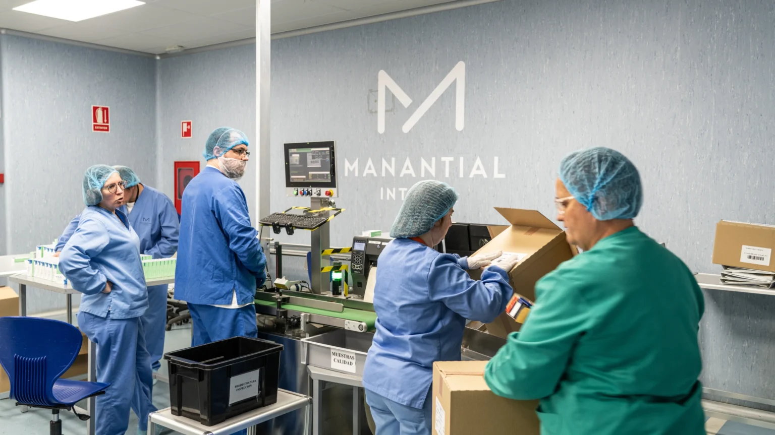 Varias personas con batas azules y verdes, redecillas para el pelo y mascarillas trabajan en una cinta transportadora en las limpias instalaciones de MANANTIAL de Rivas. Algunos empaquetan cajas, otros observan y colaboran para garantizar que los recortes no afecten a la salud mental ni a la calidad del producto.