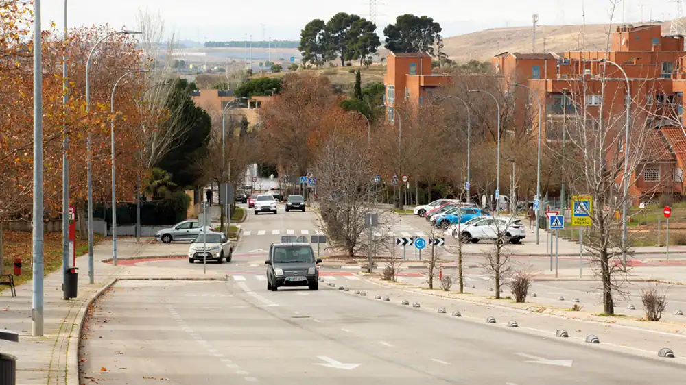Una calle con coches circulando y aparcados, rodeada de árboles sin hojas y edificios de apartamentos anaranjados. Señales de tráfico, una rotonda, pasos de peatones y colinas bajo un cielo nublado completan la escena de este borrador automático de la vida urbana.