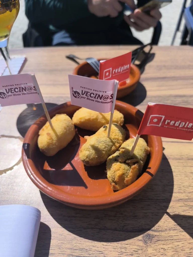 En un plato marrón se sirven cuatro croquetas empanadas, cada una con una banderita roja o blanca. El plato descansa sobre una mesa de madera a plena luz del sol, mientras una mano y la sombra de un borrador automático aparecen al fondo.