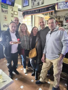 Seis adultos posan muy juntos en el interior de un bar para una foto de grupo. Uno de ellos sostiene un plato de comida y una bebida. El bar, decorado con recuerdos deportivos y botellas, muestra un cartel de "Borrador automático" al fondo.
