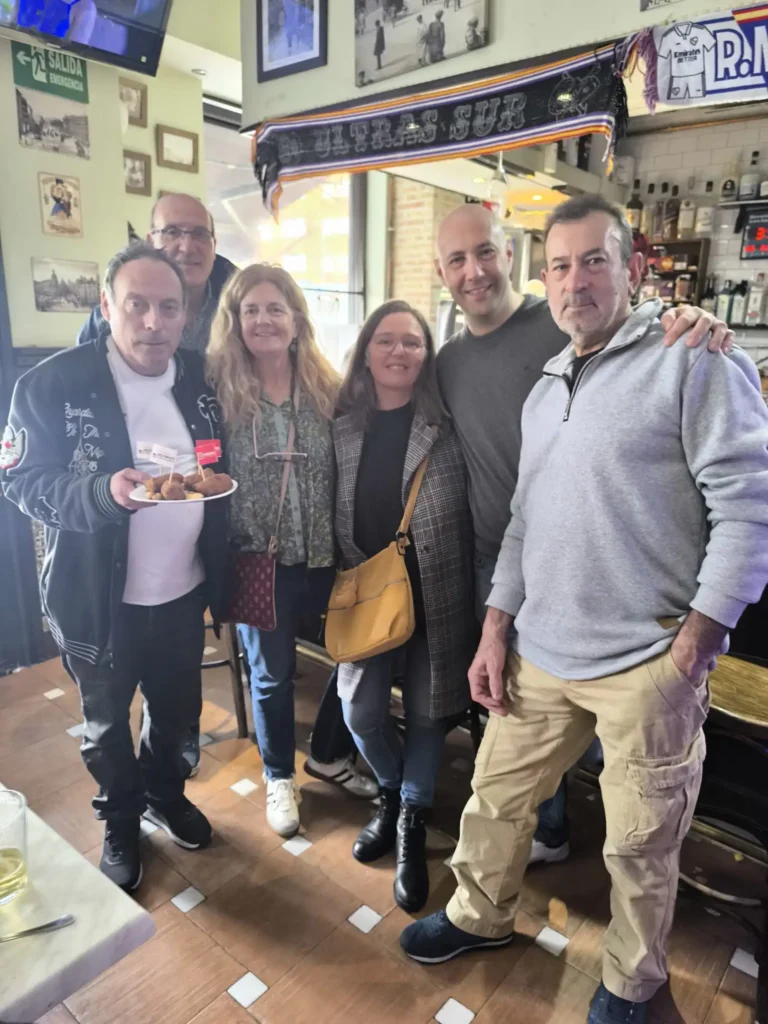 Seis adultos posan muy juntos en el interior de un bar para una foto de grupo. Uno de ellos sostiene un plato de comida y una bebida. El bar, decorado con recuerdos deportivos y botellas, muestra un cartel de "Borrador autom&aacute;tico" al fondo.