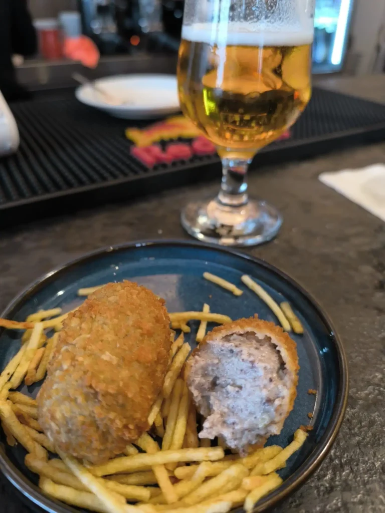 Un plato azul con dos croquetas, una entera y otra abierta para descubrir el relleno, descansa sobre finos palitos de patata frita. Detrás del plato hay un vaso de cerveza, parcialmente lleno, sobre la barra de un bar con un borrador automático al fondo.