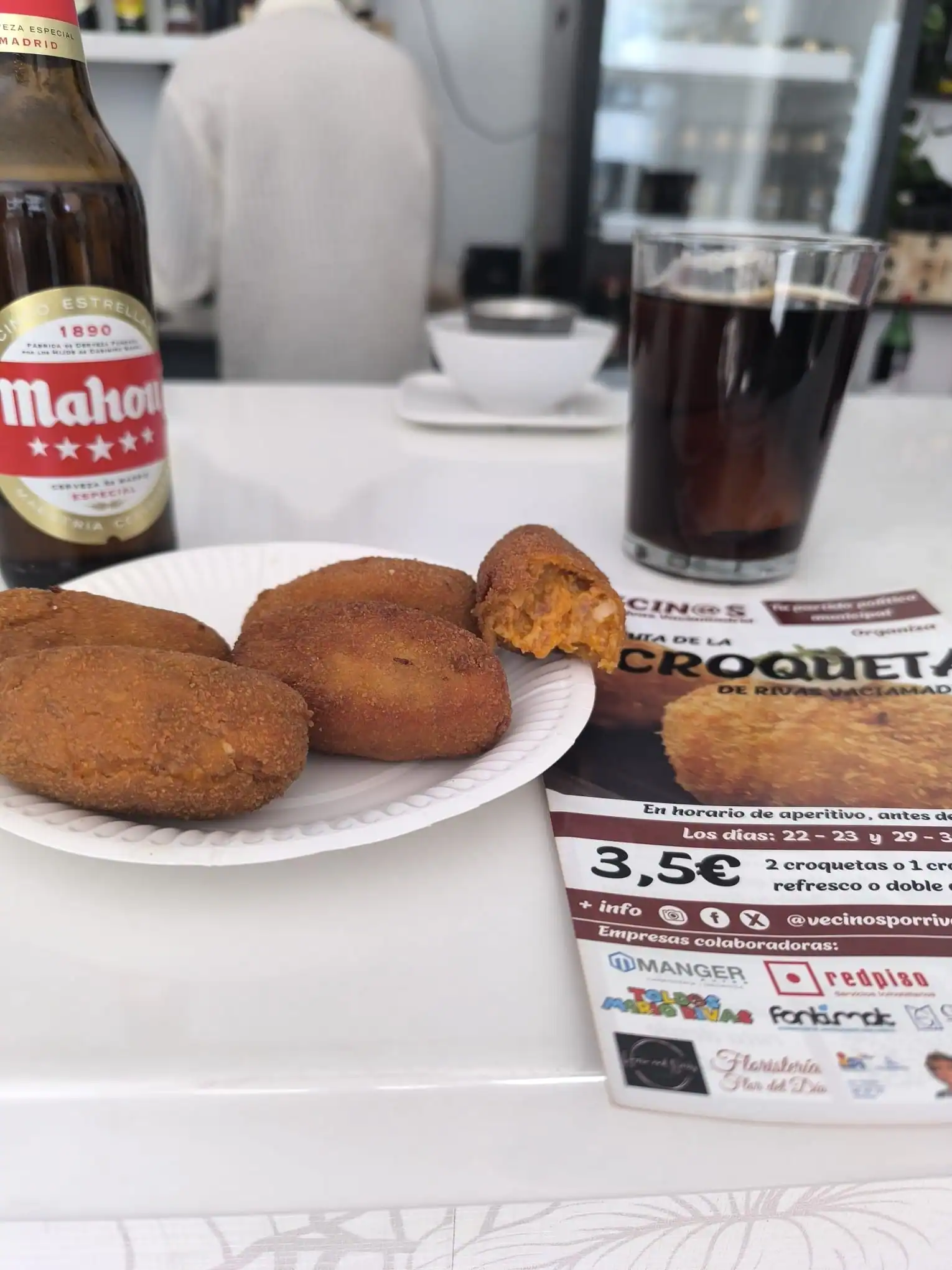 Un plato blanco con cuatro croquetas, una de ellas parcialmente comida, está sobre un mostrador junto a una botella de cerveza Mahou y un vaso de gaseosa oscura. Al fondo se ve un folleto de Borrador automático y otros objetos. Un plato blanco con cuatro croquetas, una de ellas parcialmente comida, está sobre un mostrador junto a una botella de cerveza Mahou y un vaso de gaseosa oscura. Al fondo se ve un folleto de Borrador automático y otros objetos.