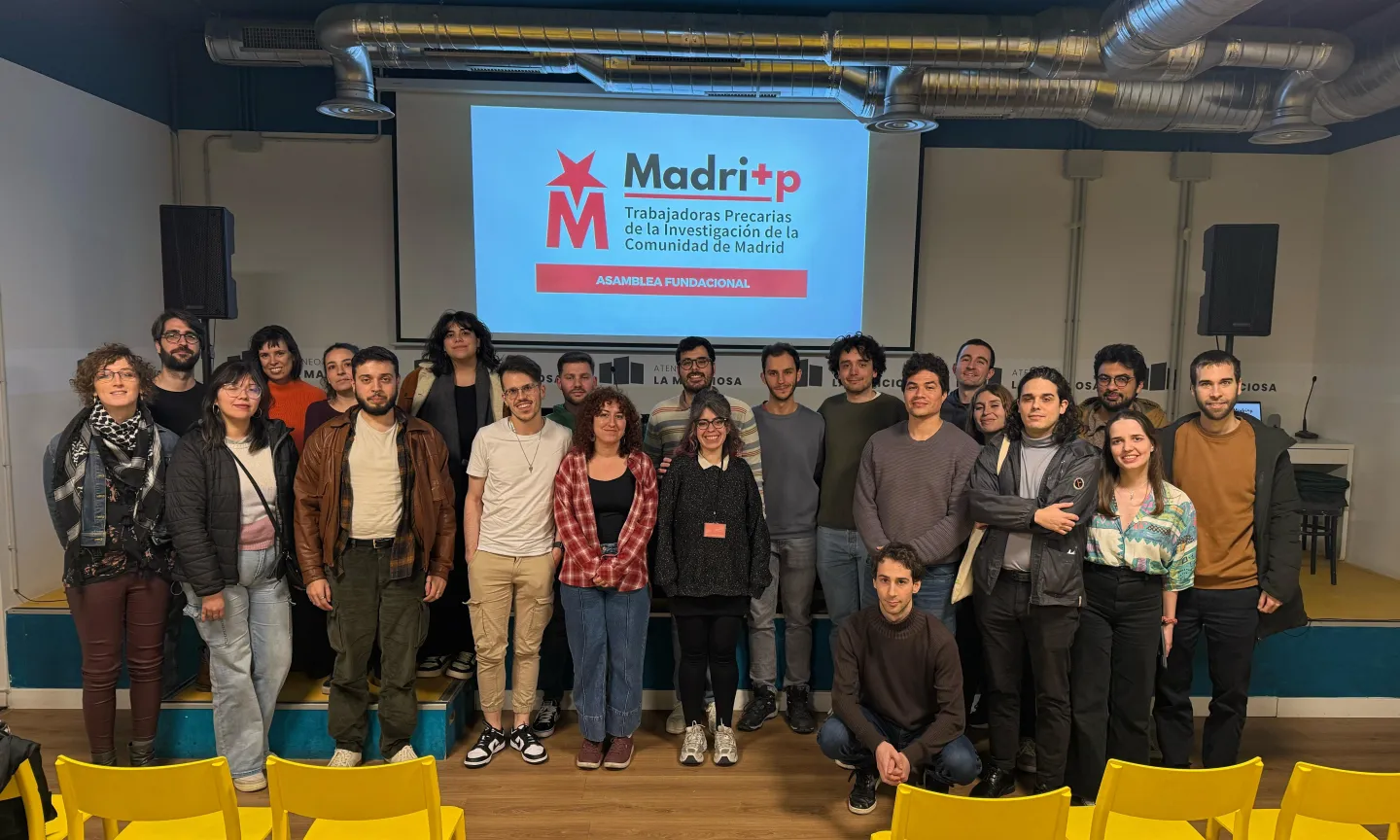 Un grupo de unas 24 personas posa para una foto de grupo delante de una pantalla en la que se ve un logotipo rojo y blanco y un texto en español sobre la asamblea de investigadores de Madrid, en la que se debate el tema del borrador automático. En primer plano se ven sillas amarillas.