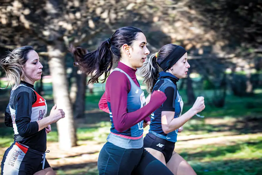 Tres mujeres corren al aire libre en el Cross de Rivas, con ropa de atletismo y dorsales. Árboles y vegetación crean un fondo difuminado, mostrando el espíritu del atletismo popular.