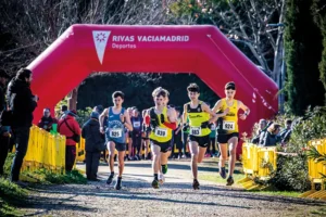 Cuatro corredores masculinos con dorsales numerados dan la salida del Cross de Rivas por un camino de grava, bajo un arco hinchable rojo rotulado Rivas Vaciamadrid Deportes, mientras los espectadores observan desde detr&aacute;s de las barreras amarillas. Una fiesta del atletismo popular.